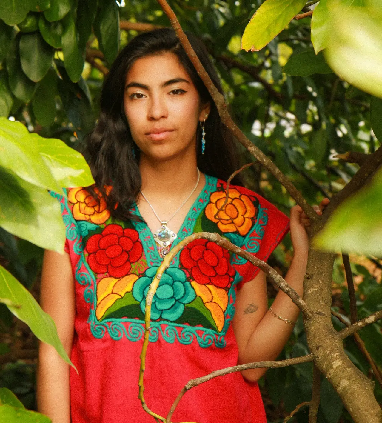 Photo: Jessika Viveros Photo of Solimar, a womanwith brown skin, hair, and eyes. She is standing with her hand leaned on an avocado tree, wearing a pink embroidered blusa, earrings from Creaciones x Solimar, and additional jewelry on her neck and wrist.